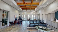 This single-floor community space makes a strong architectural statement from the moment you enter. The main lounge area commands attention with soaring ceilings adorned by a striking coffered grid of warm, natural wood beams, complemented by recessed lighting that highlights the craftsmanship throughout. Expansive arched transom windows line the front facade, flooding the interior with abundant natural light and offering views to the exterior grounds.

The flooring throughout is a rich slate tile laid in a diagonal pattern, with an ornate mosaic inlay near the entryway adding an elegant focal point. The reception and workspace area is positioned near the front, featuring a traditional wood desk and seating, framed by glass-paned French doors that connect seamlessly to adjacent rooms.

A comfortable lounge seating area anchors the main space, furnished with a full leather sofa, accent chairs in teal, and a glass-top coffee table atop layered area rugs. Opposite the seating area, a built-in entertainment wall features floor-to-ceiling gray cabinetry with granite countertops, flanking a wall-mounted television set beneath a decorative arched teal accent wall.

A bright, windowed dining nook with a round table and upholstered chairs provides a more intimate gathering space, bathed in natural light from additional arched French doors. The overall ambiance is polished, welcoming, and designed for both functional and social use.