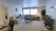 This single-floor space centers around a well-appointed living room that combines comfort and functionality. The living room is carpeted in a neutral tone, creating a warm and inviting atmosphere throughout. Large windows dressed with floral Roman shades allow generous natural light to fill the room, enhancing its open and airy feel. The space is furnished with a plush sofa accented by decorative throw pillows and a matching armchair, both in complementary neutral tones. A classic oval coffee table with cabriole legs serves as a central focal point, while a dark wood end table alongside a traditional floor lamp adds both style and practicality. An ornate wrought-iron console table positioned near the window provides additional surface space, and a wall clock adds a timeless decorative touch. Lush potted plants are strategically placed to bring a natural element into the room. The neutral wall palette and light ceiling contribute to a clean, uncluttered aesthetic, making the space feel welcoming and versatile for everyday living.