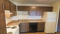 This single-floor space offers a practical and straightforward layout designed for everyday functionality. The kitchen is a central feature, showcasing dark wood cabinetry that provides ample upper and lower storage throughout. A continuous laminate countertop runs along the perimeter, offering generous workspace for meal preparation. The kitchen is equipped with essential appliances, including a white electric range, a white refrigerator, and a black built-in dishwasher seamlessly integrated beneath the counter. A single-basin sink with a standard faucet is positioned along the countertop, complemented by a dedicated shelf above for added convenience. The textured ceiling adds a classic touch to the interior, while light-toned flooring ties the space together. The overall ambiance is practical and utilitarian, with a neutral color palette of beige and warm brown tones creating a cohesive and understated aesthetic. This space is well-suited for those seeking a straightforward, move-in-ready layout with all the essential kitchen amenities in place.