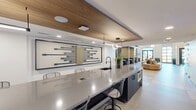 North Grove Office and Pods is a 2,280-square-foot single-floor commercial space located in Riverside, CA 92501, designed to accommodate a variety of work styles through a thoughtful blend of open and semi-private environments. The space makes a strong first impression with its soaring double-height ceilings, dramatic wood slat ceiling panels, and large floor-to-ceiling windows along the entry facade that flood the interior with natural light. Large-format neutral tile flooring runs throughout, lending a clean and cohesive aesthetic. The open lounge area features a curved sectional sofa accented with teal and gold throw pillows, centered beneath a sculptural pendant light fixture. A standout decorative wall installation composed of geometric teal panels adds an artistic focal point. Private work pods with glass-panel doors and darkened interiors provide acoustic separation for focused work or meetings. A built-in booth seating area with teal upholstered benches and wood-paneled partitions offers a semi-private collaborative nook. A dedicated kitchenette or beverage station is visible behind glass, supporting daily workplace needs. The hallway connects the various functional zones seamlessly. Overall, the space exudes a polished, contemporary ambiance suited for a professional office or coworking environment.