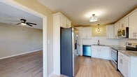 This two-story home in Fayetteville, NC offers a well-designed layout across two floors. The kitchen is a standout space, featuring white shaker-style cabinetry extending to the ceiling, granite countertops, a classic subway tile backsplash, and a full suite of stainless steel appliances including a French door refrigerator, dishwasher, microwave, and range. Light hardwood flooring and pendant lighting give the kitchen a clean, contemporary feel that flows into the adjacent living area, which is equipped with a ceiling fan. A convenient powder room on the main level includes a pedestal sink and mirror. The living areas feature light hardwood flooring throughout, while the bedrooms are finished with plush carpet and include walk-in closets with built-in shelving. The bathrooms are well-appointed, with the primary bath showcasing a white double-vanity, hardwood flooring, and stylish industrial-style lighting above a wide mirror. Natural light filters through slatted blinds in multiple rooms. The attached garage provides ample space and includes an automatic opener, electrical panel, and water heater. The overall aesthetic is neutral and move-in ready, with warm beige tones and clean white trim throughout.