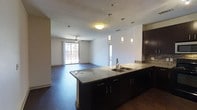 This single-floor space showcases a thoughtfully designed open-concept layout that seamlessly connects the kitchen and living areas, creating a bright and cohesive environment. The kitchen is a standout feature, boasting an expansive dark-cabinetry island topped with striking granite countertops and a double stainless steel sink. Three elegant pendant lights hang above the island, providing both functional illumination and a stylish focal point. A tiled backsplash adds texture and visual interest behind the upper cabinetry. Rich, dark hardwood flooring runs throughout the space, lending warmth and sophistication to the overall aesthetic. The open living area flows naturally from the kitchen, bathed in natural light streaming through a large sliding glass door that leads to an outdoor balcony or patio. A ceiling fan is centrally positioned in the living area, offering both comfort and airflow. The clean white walls and neutral palette create a versatile backdrop, while the combination of pendant and flush-mount ceiling lighting ensures the space is well-lit at all times. The overall ambiance is modern, clean, and inviting, with high-quality finishes throughout that reflect a contemporary design sensibility.