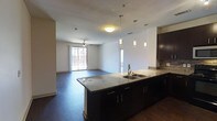 This single-floor space showcases a thoughtfully designed open-concept layout that seamlessly connects the kitchen and living areas, creating a bright and cohesive environment. The kitchen is a standout feature, boasting an expansive dark-cabinetry island topped with striking granite countertops and a double stainless steel sink. Three elegant pendant lights hang above the island, providing both functional illumination and a stylish focal point. A tiled backsplash adds texture and visual interest behind the upper cabinetry. Rich, dark hardwood flooring runs throughout the space, lending warmth and sophistication to the overall aesthetic. The open living area flows naturally from the kitchen, bathed in natural light streaming through a large sliding glass door that leads to an outdoor balcony or patio. A ceiling fan is centrally positioned in the living area, offering both comfort and airflow. The clean white walls and neutral palette create a versatile backdrop, while the combination of pendant and flush-mount ceiling lighting ensures the space is well-lit at all times. The overall ambiance is modern, clean, and inviting, with high-quality finishes throughout that reflect a contemporary design sensibility.
