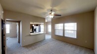 This single-floor residence offers a practical and comfortable open-concept layout designed to maximize both functionality and livability. The living area is spacious and carpeted, providing a warm, cozy feel underfoot, while a ceiling fan with an integrated light fixture keeps the space comfortable year-round. Large windows dressed with horizontal blinds line the walls, allowing generous amounts of natural light to fill the room and create a bright, airy ambiance throughout the day.

A standout feature of this space is the pass-through kitchen, which connects seamlessly to the living area through a wide opening fitted with a countertop bar — ideal for casual dining or entertaining. The kitchen is well-equipped, featuring dark wood cabinetry, a stainless steel refrigerator with an ice and water dispenser, and a sink positioned at the pass-through counter. Pendant lighting above the kitchen area adds a decorative touch and enhances task lighting. The open sightlines between the living and kitchen areas foster a connected, sociable atmosphere throughout the home. Overall, the space balances a neutral color palette with practical design elements, offering a clean and inviting environment.