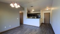 This single-floor space showcases an open-concept layout that integrates the living area and kitchen into one cohesive, functional environment. The kitchen is outfitted with dark espresso cabinetry, granite countertops, and black appliances, including a refrigerator, stove, and built-in microwave. A peninsula-style breakfast bar defines the boundary between the kitchen and the living space, offering both additional prep space and a casual dining option. Pendant lighting above the peninsula provides focused task illumination, complementing the chandelier positioned in the adjacent living area, which adds a touch of elegance. The living area is generously proportioned, featuring durable hardwood-style flooring in a rich, dark tone that extends throughout the visible space, creating a warm and unified aesthetic. Neutral gray walls provide a versatile backdrop adaptable to various décor styles. Two doors are visible—one likely leading to an entrance and another to an interior room—suggesting a practical, well-organized floor plan. The raised ceiling height contributes to an open, airy atmosphere throughout the space.