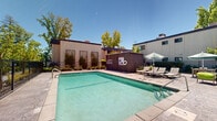 Located at 4729 Manzanita Ave in Carmichael, California, this outdoor amenity space centers around a generously sized, crystal-clear swimming pool that serves as the focal point of the area. The pool is framed by a wide concrete surround that provides ample space for lounging and movement, with multiple chaise lounge chairs and patio seating arranged throughout the deck. Shade umbrellas and a covered pergola structure with seating offer comfortable retreats from the California sun, making the space functional across all hours of the day. Vibrant green accent chairs add a pop of color against the neutral hardscape tones. The area is enclosed by a secure metal fence, ensuring safety and privacy, while mature trees surrounding the perimeter create a natural, shaded backdrop. Manicured landscaping with planters and shrubs lines the boundaries of the space, enhancing its visual appeal. A dedicated utility and facilities building is positioned on-site, complete with posted pool rules and safety signage. The multi-story residential buildings visible in the background provide context for the community setting. Under clear blue skies, this outdoor space delivers a resort-like atmosphere well-suited for relaxation and recreation.
