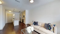 This well-designed single-floor residence offers a seamless open-concept layout that connects the living and kitchen areas, creating a cohesive and functional living environment. The living area is bright and inviting, featuring rich dark hardwood flooring that flows throughout the space and crisp white walls that enhance the sense of openness. A ceiling fan with a flush-mounted light fixture provides both comfort and illumination. The kitchen is thoughtfully designed with cream-toned cabinetry, stainless steel appliances including a refrigerator, microwave, and range, and a peninsula with pendant lighting overhead, offering both prep space and a casual dining option. A sleek sink with modern fixtures completes the kitchen. Track lighting in the kitchen ensures excellent task lighting throughout. The clean, neutral color palette runs consistently across both spaces, lending a polished and cohesive aesthetic. The combination of warm wood flooring, white walls, and stainless steel accents gives this residence a contemporary yet welcoming ambiance, well-suited for both everyday living and entertaining.