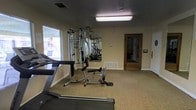 This single-floor fitness room is designed to provide a functional and practical workout environment. The space is outfitted with a multi-station cable weight machine that supports a wide range of strength training exercises, complemented by cardio equipment including a treadmill. A large wall-mounted mirror expands the visual depth of the room while allowing users to monitor their form during exercise. The flooring features neutral-toned carpet, providing a comfortable surface underfoot. Natural light filters in through a window fitted with horizontal blinds, offering a pleasant connection to the outdoors. Overhead, a fluorescent ceiling fixture ensures the room is well-lit throughout. A textured ceiling adds a traditional architectural touch, while the warm beige walls contribute to a clean and focused atmosphere. A floor-mounted ventilation register provides climate control, keeping the environment comfortable during workouts. A wooden-framed doorway leads to an adjacent corridor, offering convenient access to the rest of the facility. Overall, this fitness room is a straightforward, purpose-built space suited for residents or occupants seeking a convenient on-site exercise option.