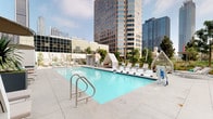 This rooftop amenity deck offers an impressive outdoor retreat elevated above a bustling urban downtown. At its centerpiece is a generously sized rectangular swimming pool with crystal-clear turquoise water, finished with sleek tile detailing and accessible via stainless steel entry rails. The surrounding pool deck is paved in large-format light gray stone tiles, providing ample space for lounging and circulation.

Lining the pool are rows of white chaise lounges accented with teal cushions, offering plentiful relaxation options in the open air. A standout feature of the deck is its collection of distinctive tent-style private cabanas with white canopies and sheer drapery, each furnished with cushioned seating and tropical-patterned pillows, providing shaded retreats with sweeping city views.

The perimeter of the deck is thoughtfully landscaped with large decorative planters, drought-tolerant plants, ornamental grasses, succulents, and slender trees, creating a lush contrast against the surrounding high-rise skyline. Outdoor shower facilities and additional seating areas are also available on deck.

The entire space is framed by panoramic views of gleaming glass towers, lending the area a sophisticated, resort-like atmosphere within the heart of the city. The combination of modern design elements, verdant greenery, and open sky creates an exceptional urban oasis.