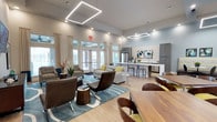 This single-floor space is a thoughtfully designed communal lounge that seamlessly blends comfort and contemporary style. The open-concept layout creates a spacious, airy atmosphere, with soaring ceilings and geometric linear pendant lighting serving as striking focal points overhead. Large French doors and floor-to-ceiling windows line one wall, flooding the interior with natural light and offering views of the outdoor surroundings.

The lounge area is furnished with an eclectic yet cohesive mix of seating, including patterned accent chairs, a plush sofa with vibrant throw pillows, and sleek modern chairs arranged over a blue area rug with abstract detailing. Wood-accented side tables and statement pieces add warmth to the otherwise light and neutral color palette.

A full-service kitchen bar anchors the far end of the space, featuring a long white countertop with bar-height seating, a stainless steel refrigerator, and a decorative tile backsplash. Gray cabinetry provides ample storage while adding a refined, modern touch. A dining area is also present, complete with rich wooden furniture that complements the light hardwood-style flooring throughout. The overall ambiance is welcoming, polished, and well-suited for both social gatherings and relaxed everyday use.
