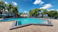 Located at 518 Lake Bridge Ln in Apopka, Florida, this outdoor amenity area centers around a generously sized swimming pool with clear, blue-green water. The pool features a defined shallow entry point and is framed by a broad, well-maintained paver deck that provides ample space for relaxation and recreation. Surrounding the pool, an array of blue lounge chairs and shaded umbrella tables are thoughtfully arranged, offering residents comfortable spots to unwind in the Florida sunshine. A dedicated outdoor dining area with a striped canopy umbrella and seating for multiple guests adds a social gathering element to the space. The pool area is enclosed by a black wrought-iron fence, providing both security and a clean aesthetic boundary. Beyond the fence, a serene pond with a decorative fountain enhances the natural ambiance, while mature trees — including palms and large shade trees — frame the perimeter, creating a lush, private retreat. The surrounding residential buildings feature traditional architectural details with neutral-toned exteriors and pitched rooflines. A single-story clubhouse or amenity building sits adjacent to the pool, adding functional convenience to the outdoor space. The overall atmosphere is vibrant, relaxing, and distinctly Florida in character.