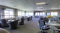 This single-floor commercial space offers a well-appointed and versatile environment suited for professional gatherings, corporate events, or collaborative work settings. The open layout is defined by a drop-panel ceiling with recessed and fluorescent lighting, complemented by generous natural light streaming in through large windows that line multiple walls, creating a bright and welcoming atmosphere throughout.

The seating arrangement is thoughtfully varied, featuring clusters of patterned upholstered lounge chairs that encourage informal conversation and relaxed collaboration. Toward one side of the space, tall bistro-style tables with bar stools provide an alternative setting for standing or elevated seating, while standard office task chairs and work surfaces are also present, offering flexibility for different uses.

Neutral-toned carpeting covers the floors throughout, adding warmth and acoustic comfort to the room. A tall decorative plant near the window area introduces a touch of greenery, softening the otherwise professional aesthetic. The overall color palette—comprising grays, blues, and muted earth tones—lends the space a polished yet approachable character. With its abundance of natural light, flexible furniture configurations, and clean design, this space is well-suited to serve as a lounge, meeting area, or multi-purpose event venue.