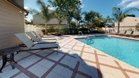 This inviting outdoor pool area is designed with both comfort and functionality in mind. The expansive pool deck is finished with a distinctive patterned tile layout — large light-colored square tiles accented by diagonal terracotta-toned border tiles — creating an elegant and visually striking surface throughout the space. The sparkling swimming pool takes center stage, offering clear blue water and step entry, surrounded by ample deck space for lounging and socializing. Multiple sling-style chaise lounges are arranged along the poolside, complemented by a large shade umbrella, providing plenty of options for sun or shade. The pool area is enclosed by white fencing, offering a sense of privacy while maintaining an open, airy feel. Lush tropical landscaping, including tall palm trees and mature greenery, frames the perimeter, enhancing the resort-like atmosphere. An adjacent building with taupe lap siding, large windows, and glass French doors opens directly onto the pool deck, providing convenient interior access. Safety equipment, including a life ring, is mounted on the building exterior. The entire setting benefits from abundant natural light under open skies, creating a bright and welcoming outdoor retreat suited for leisurely enjoyment.