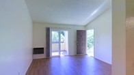 This single-floor space offers a clean, functional layout with an open and airy feel. The main living area is finished with rich hardwood-style flooring that runs throughout, complementing the neutral, light-toned walls and providing a warm, inviting atmosphere. A textured popcorn ceiling adds a classic touch overhead.

Natural light fills the room generously through a large sliding glass door that opens directly onto a private balcony, offering pleasant views of mature, green trees. A secondary entrance door with a traditional six-panel design is positioned nearby, with an adjacent sidelight that further brightens the interior. The combination of these openings creates excellent cross-lighting and a strong connection to the outdoors.

A wall-mounted heating unit is conveniently positioned along one wall, providing practical climate control without occupying floor space. The overall layout is straightforward and versatile, well-suited for a variety of furniture arrangements. The space benefits from its single-story configuration, making it easily accessible. With its natural light, outdoor access, and functional finishes, this space presents a comfortable and practical living environment.