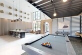 Located at 75 NE Sixth Ave in Delray Beach, Florida, this upscale amenity clubhouse delivers an impressive blend of modern design and resort-style comfort across a single, expansive floor. The grand entrance hall welcomes guests with soaring double-height ceilings, large-format light tile flooring, and sculptural pendant light fixtures that set an elegant tone throughout. The main living area opens into a voluminous, light-filled social hub anchored by a billiards table and foosball station, separated from the rest of the space by sleek vertical slatted partitions and an exposed black steel ceiling grid. A dedicated lounge zone features a refined coffered ceiling with warm wood inlays, rattan-framed seating, a patterned geometric floor, and a full wet bar with dark cabinetry and decorative tile backsplash. A communal dining area accommodates a long shared table beneath woven pendant lighting, framed by a textured wood slat accent wall adorned with organic stone wall art. Floor-to-ceiling glass doors throughout flood the interior with natural light and provide seamless access to lush tropical outdoor areas with palm trees and a pool terrace. A curated bar nook with backlit shelving and an additional lounge with styled bookshelves complete this thoughtfully appointed gathering space.
