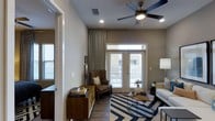 This single-floor residence offers a thoughtfully designed living area that balances comfort and style. The main living space is anchored by a ceiling fan with integrated lighting, recessed ceiling lights, and neutral-toned walls that create a calm, welcoming ambiance. Rich hardwood-style flooring runs throughout, adding warmth and visual continuity to the space.

A standout feature is the set of French doors fitted with transom windows above, flooding the room with natural light and providing direct access to a private outdoor balcony. Flowing drape panels frame the doors elegantly, adding a refined touch to the space.

The living area is generously proportioned, accommodating a full seating arrangement that includes a sofa, a classic wingback armchair, and a geometric-patterned area rug in bold navy and white tones that anchors the room. A mid-century style sideboard adds functional storage while contributing to the overall aesthetic.

The layout also incorporates a transitional entryway, suggesting a well-organized floor plan with distinct zones for living and entry. Natural light from multiple window sources enhances the sense of openness throughout. Overall, the space combines practical design with curated décor elements to deliver a comfortable and visually appealing single-floor home.