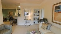 This single-floor residence offers a well-appointed living area finished in soft, neutral tones that create a warm and inviting atmosphere. The living space is carpeted and features recessed lighting alongside crown molding details that lend a polished architectural touch to the room. A standout element is the built-in shelving unit along one wall, providing both display and storage space, complemented by an adjacent door for added functionality. A decorative console table with an elegant wrought-iron base serves as a focal point along the central wall, accented by framed artwork and a small table lamp that contribute to the room's refined aesthetic. Natural light filters into the space through a pass-through opening that connects the living area to the kitchen, where bar-style seating along a raised counter creates a casual dining option. The kitchen area features green accent walls that contrast neatly with the otherwise neutral palette. Lush indoor plants are positioned throughout the living space, adding organic texture and life to the interior. Overall, the layout emphasizes an open, connected flow between the living and kitchen areas, making the space feel airy and functional.