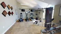 This fitness center offers a thoughtfully designed workout environment that balances functionality with an inviting aesthetic. The space features soaring vaulted ceilings that contribute to an open, airy atmosphere, while striking arched windows allow generous amounts of natural light to pour in throughout the day. Neutral sage-green walls and patterned carpet flooring create a calm, focused setting for exercise.

The cardio zone is well-appointed with multiple treadmills, elliptical trainers, and a stationary bike, each equipped with individual screens for entertainment. Wall-mounted flat-screen televisions are positioned for easy viewing from the cardio equipment. The strength training area includes a multi-function cable machine, a weight bench, free weights, and additional resistance equipment to support a full-body workout routine.

Decorative framed artwork and geometric wall accents add a polished, finished look to the space. Overhead linear fluorescent lighting supplements the abundant natural light on overcast days. A welcoming entry area features recessed lighting, double glass doors, and a seating bench, providing a clean transition into the facility. An adjacent room visible through interior glass doors suggests additional amenity spaces nearby. Overall, this fitness center is both practical and visually appealing.