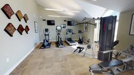 This fitness center offers a thoughtfully designed workout environment that balances functionality with an inviting aesthetic. The space features soaring vaulted ceilings that contribute to an open, airy atmosphere, while striking arched windows allow generous amounts of natural light to pour in throughout the day. Neutral sage-green walls and patterned carpet flooring create a calm, focused setting for exercise.

The cardio zone is well-appointed with multiple treadmills, elliptical trainers, and a stationary bike, each equipped with individual screens for entertainment. Wall-mounted flat-screen televisions are positioned for easy viewing from the cardio equipment. The strength training area includes a multi-function cable machine, a weight bench, free weights, and additional resistance equipment to support a full-body workout routine.

Decorative framed artwork and geometric wall accents add a polished, finished look to the space. Overhead linear fluorescent lighting supplements the abundant natural light on overcast days. A welcoming entry area features recessed lighting, double glass doors, and a seating bench, providing a clean transition into the facility. An adjacent room visible through interior glass doors suggests additional amenity spaces nearby. Overall, this fitness center is both practical and visually appealing.