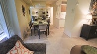 This single-floor residence offers a thoughtfully designed open-concept layout that promotes a seamless flow between the living, dining, and kitchen areas. The dining space is centrally positioned and comfortably accommodates a full dining table with seating for multiple guests, making it ideal for both everyday meals and entertaining. A large mirror on the dining room wall enhances the sense of space and reflects warm ambient lighting throughout the room.

Soft, neutral-toned carpeting extends across the living and dining areas, contributing to a warm and inviting atmosphere. Decorative wall art adds character to the space, while understated, elegant furnishings maintain a clean and cohesive aesthetic.

The kitchen is partially open to the dining area via a pass-through counter framed by a decorative lattice-style divider, creating a visual connection between spaces while maintaining a sense of definition. The kitchen features light cabinetry, including warm wood-toned upper cabinets, and is finished with clean, light-colored surfaces that complement the overall palette of the home.

Recessed lighting illuminates the interior evenly, and the neutral color scheme throughout creates a calm, functional, and welcoming environment suitable for a variety of living needs.