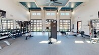 This fitness center offers an expansive, single-floor workout space designed to accommodate a wide range of training styles. The facility's most striking architectural feature is its trio of full-height glass panel garage doors along one wall, which flood the interior with abundant natural light and provide open views of the outdoor greenspace beyond. Clerestory windows positioned above the garage doors further enhance the bright, airy atmosphere throughout the space.

The soaring ceiling height adds a sense of grandeur and openness, and is accented by a coffered teal ceiling detail at the center, from which a large industrial ceiling fan is suspended — contributing to both the aesthetic and comfort of the space. The walls feature a bold contrast of white and warm orange tones, adding energy and visual interest to the environment.

Flooring transitions between durable hardwood-style planks and commercial-grade carpet tile, delineating functional zones within the gym. The equipment selection is comprehensive, including cable machines, free weights, weight benches, treadmills, ellipticals, a StairMaster, and functional training implements such as a tire flip station. Large mirrors along one wall support strength training and form checks. The combination of thoughtful design and diverse equipment makes this fitness center a well-rounded facility suited for a full spectrum of workout routines.