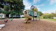 This outdoor recreational space centers around a well-equipped playground structure designed to engage children of various ages. The standout feature is a multi-level play unit constructed with a durable metal and molded plastic framework in neutral tan and teal tones. A prominent spiral slide serves as the primary descending element, twisting downward from an elevated platform to ground level. The structure also incorporates a climbing tower with ladder access, interactive panel activities, and connecting platforms that encourage exploration and physical activity.

The ground surface beneath and surrounding the play equipment is covered with rubber mulch, providing a safety-conscious, impact-absorbing layer appropriate for active play. The area is generously shaded by mature deciduous trees, whose broad canopies offer natural relief from direct sunlight and contribute to an inviting outdoor environment. The backdrop includes a multi-story residential building with large windows and well-maintained landscaping, integrating the playground naturally into the surrounding community setting. Neatly trimmed shrubs and green lawn areas frame the perimeter, adding a polished, cared-for appearance to the space overall.