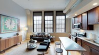 This single-floor loft-style space showcases a compelling blend of industrial character and contemporary design. The open-concept layout connects the living and kitchen areas in a seamless, functional arrangement that maximizes both space and versatility.

The living area is anchored by a set of striking, oversized factory-style windows with bold black frames, allowing an abundance of natural light to flood the interior while offering serene views of surrounding bare-branch trees. The exposed ductwork on the ceiling and light-toned hardwood flooring reinforce the industrial aesthetic, while the warm lighting and carefully selected furnishings — including a dark sofa, sleek accent chairs, and a glass coffee table — lend a modern, refined feel.

Adjacent to the living space, the kitchen features rich dark cabinetry, stainless steel appliances, and a clean white breakfast bar with bar stools, providing a practical and stylish dining option. The kitchen is efficiently laid out, making the most of the available space.

Throughout, the interplay of raw industrial elements — exposed brick accents, visible ductwork, and black-framed windows — with polished modern finishes creates a distinctive, urban ambiance that is both visually engaging and highly livable.