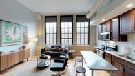 This single-floor loft-style space showcases a compelling blend of industrial character and contemporary design. The open-concept layout connects the living and kitchen areas in a seamless, functional arrangement that maximizes both space and versatility.

The living area is anchored by a set of striking, oversized factory-style windows with bold black frames, allowing an abundance of natural light to flood the interior while offering serene views of surrounding bare-branch trees. The exposed ductwork on the ceiling and light-toned hardwood flooring reinforce the industrial aesthetic, while the warm lighting and carefully selected furnishings — including a dark sofa, sleek accent chairs, and a glass coffee table — lend a modern, refined feel.

Adjacent to the living space, the kitchen features rich dark cabinetry, stainless steel appliances, and a clean white breakfast bar with bar stools, providing a practical and stylish dining option. The kitchen is efficiently laid out, making the most of the available space.

Throughout, the interplay of raw industrial elements — exposed brick accents, visible ductwork, and black-framed windows — with polished modern finishes creates a distinctive, urban ambiance that is both visually engaging and highly livable.