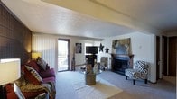 This single-floor space offers a warm and characterful living environment anchored by a standout wood-mantled fireplace that serves as the focal point of the main living area. The room features a textured ceiling with a subtle tray detail, adding architectural interest overhead, while plush carpeting underfoot contributes to a comfortable, welcoming atmosphere. Natural light filters in through vertical blinds flanking a sliding glass door, which opens to an outdoor patio area, providing a seamless connection to the exterior. The living space is generously proportioned, offering ample room for seating arrangements centered around a glass-topped coffee table. The interior is enriched by bold, eclectic styling — zebra-print accent chairs, animal-themed artwork, and mixed-pattern textiles lend the space a distinctive, lively personality. A wall-mounted television and decorative metal star accents round out the living area's design. The overall ambiance balances casual comfort with expressive, nature-inspired décor, making it a visually engaging and livable space throughout.