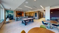 The Six Points Bloomfield Club Room is a thoughtfully designed communal amenity space located in Bloomfield, NJ. The single-floor layout encompasses several distinct functional zones, each finished with contemporary materials and modern design sensibilities throughout.

Upon entry, a sleek corridor lined with textured, striated wall panels leads past elevator access and glass-door entries, setting a clean and polished first impression. Recessed and linear LED lighting throughout ensures consistent, well-distributed illumination across all areas.

The main club room is expansive and multifunctional. A central pool table anchors the game area, complemented by a fully appointed bar counter with bar seating and a decorative reclaimed wood accent wall with pendant lighting and a mounted television. Built-in shelving adds both style and practicality to the surrounding space.

An adjacent dining and gathering area features a long, sleek communal table beneath a striking cluster of modern pendant lights, with wine refrigeration built into the cabinetry nearby. Large windows frame views of the Bloomfield streetscape, flooding the space with natural light.

A dedicated lounge area provides a relaxed retreat with modular sectional seating, accent chairs in vibrant tones, and a rich wood feature wall with integrated cove lighting. An upper mezzanine level with glass railings offers additional seating with sweeping views of the neighborhood skyline, enhanced by a sculptural ceiling installation overhead.