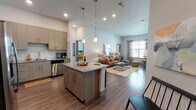 This well-appointed single-floor residence offers a thoughtfully designed open-concept layout that seamlessly connects the kitchen, dining, and living areas. The kitchen is a standout, featuring flat-front cabinetry in a warm taupe finish, granite countertops, a subway tile backsplash, and a full suite of stainless steel appliances including a refrigerator, range, microwave, and dishwasher. A center island with pendant lighting above provides additional prep space and casual dining. Rich wood-look flooring flows through the main living areas, while the spacious living room is bathed in natural light from multiple windows dressed with sheer curtains and blinds. Elevated ceilings add to the sense of openness throughout. The primary bedroom includes a walk-in closet and an en-suite bathroom with a granite-topped double vanity and a separate shower. The secondary bedroom features sliding glass doors that open directly onto a private balcony. A second full bathroom offers a tub-shower combination with tile surround, granite vanity, and large mirror. Clean lines, neutral tones, and quality finishes throughout give this residence a polished, contemporary feel.