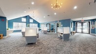 Located at 900 Crest Ln in Fort Lee, NJ, this single-floor amenity and co-working space combines functional design with a distinctive contemporary aesthetic. The interior is unified by a bold blue color palette across the walls, complemented by patterned carpet flooring that runs throughout the space.

The main open area features soaring vaulted ceilings and is anchored by two striking sputnik-style chandeliers that add a mid-century modern flair. Semi-private work pods with high white panels are arranged throughout, offering individual workstations suited for focused tasks. A dedicated workroom provides a built-in desk, seating for two, and generous natural light from oversized windows that extend up toward the roofline, framing views of the surrounding neighborhood.

A glass-door conference room is outfitted with a long table and task chairs, featuring a dramatic nature-scene mural across one wall. A separate lounge area showcases an eye-catching fan-patterned wallcovering adorned with circular gold-framed mirrors, paired with upholstered seating and a bench. Additional amenities include a reception-style counter, a utility room, and ample recessed lighting throughout, creating a well-lit, professional, and welcoming environment.