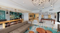 Located at 110 N Myers St in Oceanside, California, this single-floor amenity space offers a thoughtfully designed open-plan layout that blends social, recreational, and lounge functions seamlessly. The interior is defined by exposed wood beam ceiling details, light hardwood-style flooring, and white walls that create a bright, airy atmosphere throughout.

The kitchen is a standout feature, showcasing a bold teal subway tile backsplash that extends the full height of the wall, complemented by dark cabinetry, open wood shelving, stainless steel appliances including a gas range and French door refrigerator, and a large wood-topped island with bar seating. Three sculptural black pendant lights hang overhead, adding a dramatic contrast to the warm wood tones.

The main lounge area is furnished with comfortable sofas, accent chairs, and layered rugs in teal and gray tones. A sculptural multi-bulb chandelier anchors the space visually. A billiards table anchors the game area, set against a marble-effect feature wall. A separate, more intimate lounge nook features built-in light wood cabinetry with an integrated television and natural window views. Large glass doors open directly to an outdoor terrace, enhancing the connection between indoor and outdoor living.