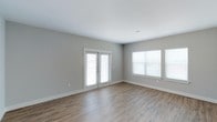 This single-floor space offers a clean, modern aesthetic defined by its understated yet functional design. The main living area is finished with warm-toned, wood-look hard flooring that runs throughout, providing both durability and visual continuity. Walls are painted in a soft, neutral grey tone that complements the bright white ceiling and crisp white baseboards, creating a light and airy atmosphere throughout.

Natural light is a standout feature of this space. A set of white-framed French doors allows abundant light to pour in while also providing direct access to the exterior. Flanking the room, additional windows fitted with white horizontal blinds further enhance the brightness and offer flexible light control. The combination of multiple light sources ensures the interior feels open and welcoming at all times of day.

The space is notably well-proportioned, offering a comfortable sense of openness without feeling oversized. The minimal detailing and neutral palette make it highly versatile, lending itself easily to a variety of furniture arrangements and interior styles. Overall, the space presents a fresh, move-in ready environment with a contemporary feel.