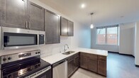 This contemporary single-floor unit offers a thoughtfully designed layout with a clean, modern aesthetic throughout. The kitchen serves as a standout feature, showcasing rich dark-toned cabinetry that extends from floor to ceiling, providing generous storage. A full suite of stainless steel appliances — including a refrigerator, electric range, built-in microwave, and dishwasher — pairs with crisp white countertops to create a stylish and functional cooking space. Pendant lighting and recessed ceiling lights add warmth and depth to the kitchen, which flows naturally into the open living area. Light-toned luxury vinyl plank flooring runs throughout the main living areas, lending a bright and cohesive feel to the space. The bedrooms are finished with neutral tones and feature large windows that invite natural light and offer views of the surrounding urban streetscape. One bedroom features plush carpeting and dark sliding closet doors, while another benefits from hard flooring and direct window access to city views. The bathroom is clean and minimalist, featuring a walk-in shower, a vanity with dark cabinetry and a white countertop, and light flooring that ties the space together. Overall, the unit presents a polished, move-in ready environment suited for modern urban living.
