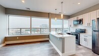Located at 2901 Euclid Ave in the heart of Cleveland, Ohio, this modern single-floor unit offers a thoughtfully designed open-concept layout with contemporary finishes throughout. The kitchen serves as a central highlight, anchored by a clean-lined island with a built-in sink and pendant lighting overhead. White shaker cabinetry, a classic subway tile backsplash, and a full suite of stainless steel appliances — including a refrigerator, electric range, and over-the-range microwave — complete the space. Expansive floor-to-ceiling windows flood the kitchen and living areas with natural light while framing elevated views of the Cleveland skyline and surrounding cityscape. Light wood-tone flooring runs seamlessly throughout, complementing the soft gray and white color palette that gives the unit a clean, airy feel. The bedroom features generous window coverage, continuing the panoramic urban views, with baseboard heating and neutral walls offering a calm, versatile space. The bathroom is ADA-accessible, finished with large-format tile flooring, a white vanity with ample drawer storage, and matte black grab bars and fixtures for a modern, inclusive design. The overall layout, as seen in the dollhouse view, is efficiently organized with clear sightlines and a functional flow throughout.