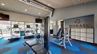 Located at 7007 E Gold Dust Ave in Scottsdale, Arizona, this fitness center offers a comprehensive, single-floor workout facility designed to accommodate a full range of exercise routines. The main workout area features eye-catching hexagonal tile flooring in bold shades of blue and gray, creating a dynamic and energizing atmosphere. The space is outfitted with multiple treadmills, elliptical machines, and a variety of strength training equipment, including cable machines, weight benches, and a full dumbbell rack. Large windows along one wall flood the cardio zone with natural light while offering views of the surrounding outdoor environment, enhancing the overall workout experience. An exposed ductwork detail and a wood-paneled ceiling add an industrial-modern aesthetic to the space, complemented by recessed lighting throughout. A separate stretching and flexibility studio features light wood-look flooring, floor-to-ceiling mirrors, and ample open floor space, stocked with exercise mats and stability balls for low-impact and functional training. Restroom facilities are conveniently accessible from this area via dark wood-finished doors. The facility is well-organized, clearly marked, and maintained to a high standard, making it a fully functional and welcoming fitness destination in the heart of Scottsdale.