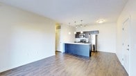This single-floor space showcases an open-concept layout that seamlessly connects the living area to the kitchen, creating a bright and airy atmosphere. The kitchen serves as the focal point, featuring dark espresso cabinetry that contrasts elegantly with light stone countertops along the peninsula island. Stainless steel appliances, including a refrigerator, microwave, and range, add a polished, contemporary feel. Above the island, a trio of pendant lights provides targeted task lighting, while a multi-bulb track light fixture illuminates the broader kitchen space. The warm-toned hardwood-style flooring runs throughout, adding continuity and depth to the open floor plan. Neutral white walls keep the space feeling clean and versatile, allowing natural and artificial light to bounce effectively across the room. A white panel door leads to an adjacent space, likely a closet or utility area, tucked neatly into the corner. The generous open living area offers flexible furniture arrangement possibilities, making the space both functional and inviting. Overall, the unit presents a well-designed, modern aesthetic with quality finishes throughout.