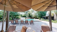Located at 3200 Hartley Rd in Jacksonville, Florida, this single-level outdoor amenity space offers a well-appointed pool and patio area designed for relaxation and social gathering. The centerpiece is a generously sized swimming pool surrounded by a broad, paver-brick deck that provides ample room for lounging and movement. Numerous sling-style chaise lounges are arranged poolside, offering residents dedicated spaces to unwind in the Florida sun.

Large shade canopies extend over a portion of the deck, providing welcome relief from the sun while maintaining an open, airy feel. Beneath the canopies, cast-iron patio tables paired with wicker and sling chairs create comfortable seating arrangements ideal for outdoor dining or casual conversation. A charcoal grill station is also available on the patio, adding to the space's outdoor entertaining appeal.

The area is enclosed by a combination of wood fencing and black metal railings, ensuring privacy and security. Mature trees and manicured hedgerows frame the perimeter, creating a lush, green backdrop that enhances the natural ambiance. A nearby building with large windows is visible from the patio, suggesting a clubhouse or leasing facility in close proximity. Overall, this outdoor space balances functionality with a relaxed, resort-inspired atmosphere.