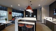 This single-floor communal space is designed with a contemporary, functional aesthetic that caters to productivity and collaboration. At its center sits a long, elevated bar-style island with a clean white surface, equipped with built-in power outlets and flanked by sleek wire-frame stools with orange upholstered seats, adding a striking pop of color against the neutral palette.

Overhead, bold red industrial pendant lights serve as a focal point, complementing the recessed ceiling lighting that provides consistent, even illumination throughout the space. The ceiling features a modern flat design with integrated ventilation, contributing to the clean, streamlined look.

Along the perimeter, multiple individual computer workstations are arranged, each equipped with monitors, making the space well-suited for focused work or research. The dark accent walls contrast with the lighter flooring and surfaces, creating a visually dynamic environment.

A lounge area with upholstered seating is positioned near floor-to-ceiling windows, offering views to the exterior and introducing natural light. The overall ambiance is polished and urban, balancing a professional atmosphere with inviting design elements.