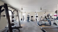 This modern fitness center is thoughtfully designed to support a wide range of workout routines in a clean, open environment. The space is bathed in natural light from large windows positioned throughout the facility, creating an energizing atmosphere that complements the crisp white walls and neutral gray carpet tile flooring.

The cardio zone is well-stocked with treadmills, elliptical machines, stationary bikes, and cycling bikes — several of which are equipped with integrated display screens for interactive training sessions. The strength training area features a robust selection of free weights, including a full dumbbell rack, adjustable benches, a power rack with barbells and weight plates, and multiple cable and resistance machines to accommodate both isolation and compound exercises.

A dedicated stretching and functional training section is defined by rubberized interlocking floor mats, providing a safe surface for floor-based exercises. Full-length wall mirrors run along key areas of the floor, helping users maintain proper form during workouts. Pendant-style cylindrical light fixtures hang from the smooth white ceiling, supplementing the generous natural light.

Practical amenities include a dual water fountain station with a bottle-filling feature, keypad-secured entry doors, and clearly posted fitness center guidelines. The overall layout is spacious and well-organized, offering a professional gym experience.
