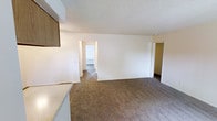 This single-floor residential space offers a straightforward, functional layout designed for comfortable everyday living. The main living area is finished with wall-to-wall neutral-toned carpet, providing a warm and soft underfoot feel throughout the space. The textured popcorn ceiling adds a classic touch and runs consistently across the living area, contributing to the room's overall character.

The open layout connects seamlessly to adjacent spaces, with clearly defined doorways leading to what appears to be a bedroom and additional rooms beyond. A hallway visible to the right provides access to further areas of the unit, enhancing the sense of connectivity throughout the floor plan.

To one side, the kitchen area is partially visible, featuring wood-toned cabinetry overhead and a light-colored countertop that contrasts neatly with the surrounding white walls. The transition from hard flooring in the kitchen to carpet in the living area creates a natural delineation between functional zones.

The crisp white walls throughout contribute to a clean, bright interior, while the overall layout reflects a practical and efficient use of space. The single-floor design ensures easy navigation with no stairs required, making the space accessible and convenient for a wide range of occupants.