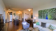 This single-floor residence offers an open-concept layout that connects the living, dining, and kitchen areas in a cohesive and functional design. The living area provides a welcoming atmosphere, furnished with a comfortable sofa, accent chair, and glass coffee table, all anchored by a patterned area rug over rich hardwood flooring that extends throughout the space. Vibrant botanical-themed artwork adds a pop of color against the clean white walls, contributing to the fresh and contemporary ambiance. The dining area sits just beyond the living space, accommodating an intimate round table and chairs, ideal for everyday meals or casual entertaining. The kitchen features a dark-toned island with bar seating, complemented by pendant lighting that casts a warm glow over the counter. The open sightlines between all areas create a sense of spaciousness and ease of movement throughout. Flush-mount and pendant light fixtures provide consistent illumination across the layout. A hallway visible from the main living area leads to additional rooms, suggesting a thoughtful and practical floor plan. The overall aesthetic is modern and inviting, with a cohesive color palette of neutrals accented by greens and teals.