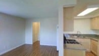This single-floor space offers a practical and functional layout designed to maximize everyday living. The main living area features warm wood-style laminate flooring that extends throughout, creating a cohesive and inviting atmosphere. A textured popcorn ceiling adds dimension overhead, while clean white walls and white trim provide a neutral backdrop adaptable to a variety of interior styles.

A doorway from the living area leads to a dedicated storage space, fitted with floor-to-ceiling built-in cabinetry with double doors, offering generous organizational capacity. A wall-mounted ventilation unit is also visible, contributing to the room's utility.

The kitchen is partially open to the living area, separated by a half-wall partition. Light wood cabinetry lines the upper portion of the kitchen, complemented by a granite-style countertop and a tiled backsplash. A gas range adds to the kitchen's functionality. The partial separation between the kitchen and living area maintains a sense of openness while defining each distinct zone. Overall, the space presents a clean, straightforward, and well-organized environment.