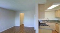 This single-floor space offers a practical and functional layout designed to maximize everyday living. The main living area features warm wood-style laminate flooring that extends throughout, creating a cohesive and inviting atmosphere. A textured popcorn ceiling adds dimension overhead, while clean white walls and white trim provide a neutral backdrop adaptable to a variety of interior styles.

A doorway from the living area leads to a dedicated storage space, fitted with floor-to-ceiling built-in cabinetry with double doors, offering generous organizational capacity. A wall-mounted ventilation unit is also visible, contributing to the room's utility.

The kitchen is partially open to the living area, separated by a half-wall partition. Light wood cabinetry lines the upper portion of the kitchen, complemented by a granite-style countertop and a tiled backsplash. A gas range adds to the kitchen's functionality. The partial separation between the kitchen and living area maintains a sense of openness while defining each distinct zone. Overall, the space presents a clean, straightforward, and well-organized environment.