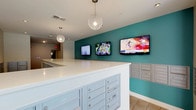 This contemporary mail room combines functionality with stylish design elements. The space is dominated by a long white counter that serves as the focal point, likely used for sorting and distributing mail. Behind the counter, a vibrant teal accent wall adds a pop of color and energy to the room. Three large flat-screen TVs are mounted on this wall, presumably to provide entertainment or information to residents while they check their mail. The room is illuminated by recessed lighting and two eye-catching spherical pendant lights, which add a touch of elegance to the utilitarian space. The ceiling appears to be standard height with a smooth finish. To the right of the counter, a bank of individual mailboxes is visible, neatly organized for easy access. The floor appears to be tiled, suggesting durability and easy maintenance. Overall, the space strikes a balance between practicality and modern aesthetics, creating an inviting atmosphere for a typically mundane task.