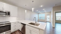 This well-appointed single-floor unit showcases a clean, contemporary design with wood-look flooring and neutral tones carried consistently throughout. The open-concept kitchen serves as a central feature, boasting crisp white shaker cabinetry, a classic subway tile backsplash, and gleaming quartz countertops. A large kitchen island with an undermount stainless steel sink and pendant lighting above offers both workspace and casual dining potential. The kitchen is fully equipped with a full suite of stainless steel appliances, including a French door refrigerator, electric range, over-the-range microwave, and dishwasher. The living areas are bright and versatile, benefiting from large windows that invite natural light and offer views of the surrounding neighborhood. The primary bathroom is a standout, featuring a generous dual-sink vanity with white cabinetry, backlit mirrors, and a walk-in glass-enclosed shower with tile surround. Additional rooms provide flexible space adaptable to bedrooms or other uses, each with ample natural light from wide windows. Recessed lighting throughout, modern hardware, and a consistent neutral color palette give the entire unit a polished, move-in-ready feel.