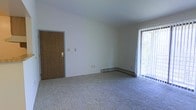 This single-floor space presents a clean, straightforward layout centered around a modestly sized living area. The room features wall-to-wall carpeting in a neutral gray tone, contributing to a quiet and comfortable atmosphere underfoot. White walls provide a blank canvas throughout, keeping the space feeling open and bright.

A prominent wood-panel interior door with a brass handle adds a warm, classic touch to the otherwise simple aesthetic. Along the far wall, a baseboard heating unit runs the length of the room, offering an efficient and unobtrusive heating solution. Light switches and electrical outlets are conveniently positioned along the walls.

Natural light is a notable feature, with a large window dressed in vertical blinds on the right side of the room. The blinds allow for flexible light control while providing a view of the greenery outside, lending a connection to the outdoors. The window's scale enhances the sense of openness within the space.

Overall, the unit maintains a functional and no-frills character, well-suited for those seeking a straightforward, comfortable living environment with practical amenities already in place.