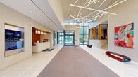 Located at 450 N 18th St in Philadelphia's vibrant Art Museum neighborhood, this grand lobby makes an immediate impression with its generous open floor plan and soaring ceiling heights. Floor-to-ceiling windows along the entrance flood the space with abundant natural light, creating a bright and welcoming atmosphere. A dramatic, sculptural chandelier composed of intersecting linear light fixtures serves as a bold centerpiece overhead, casting a warm, modern glow across the entire area. The reception desk is elegantly framed by warm wood vertical slat paneling, accented by pendant lighting that adds depth and sophistication to the feature wall. Large-format white tile flooring transitions seamlessly to carpet, defining distinct zones within the expansive hallway. Curated contemporary artwork adorns the walls, adding vibrant color and character to the otherwise refined, neutral palette of cream stone-clad walls. Sculptural red-and-black benches offer stylish seating throughout. An integrated digital display near the reception area provides informational content for visitors, while an elevator and staircase ensure smooth circulation. The space conveys a polished, professional atmosphere with a strong architectural identity, well-suited for a commercial or institutional setting.