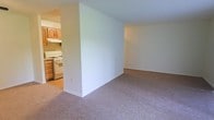 This single-floor residence offers a straightforward, functional layout that makes efficient use of its space. The living area is finished with wall-to-wall neutral-toned carpet, providing a comfortable and cohesive feel underfoot. Smooth, light-colored walls keep the space bright and adaptable to a variety of furnishing styles. A partial dividing wall separates the living area from the kitchen, creating a subtle distinction between the two spaces while maintaining an open connection.

The kitchen features warm wood-toned cabinetry with overhead and base units providing ample storage. A white gas range with an overhead ventilation hood serves as the primary cooking appliance. The kitchen flooring transitions to a hard surface, contrasting with the carpeted living area. The cabinetry and appliances reflect a classic, utilitarian design suited for everyday use.

Overall, the space presents a clean, neutral palette throughout, with minimal architectural ornamentation. The layout prioritizes practicality, with clearly defined functional zones and an uncluttered interior that offers a blank canvas for personalization.